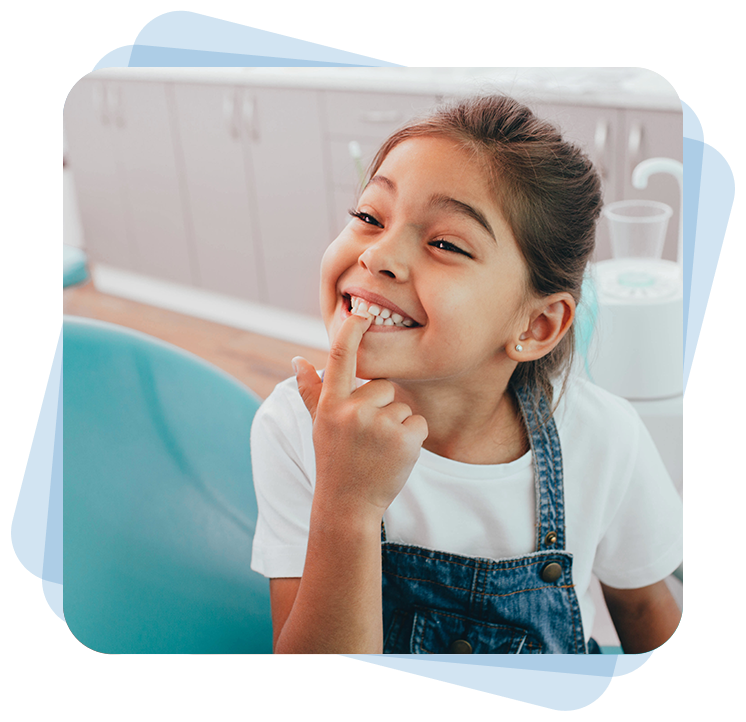 A young girl in a white shirt and denim overalls smiles widely, pointing at her mouth, sitting in a dental office.